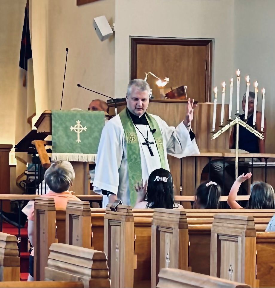 A clergy member wearing a green stole stands in a church, gesturing to children sitting in pews near a lit candelabra.