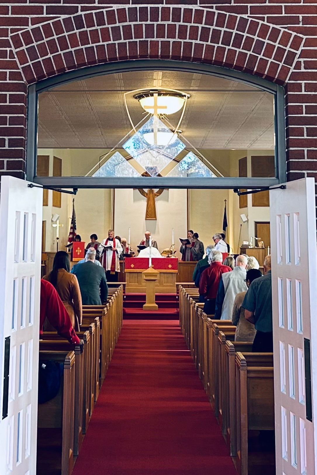 A view through open white doors into a church with a red carpet aisle, pews, and a sanctuary with a cross at the altar.