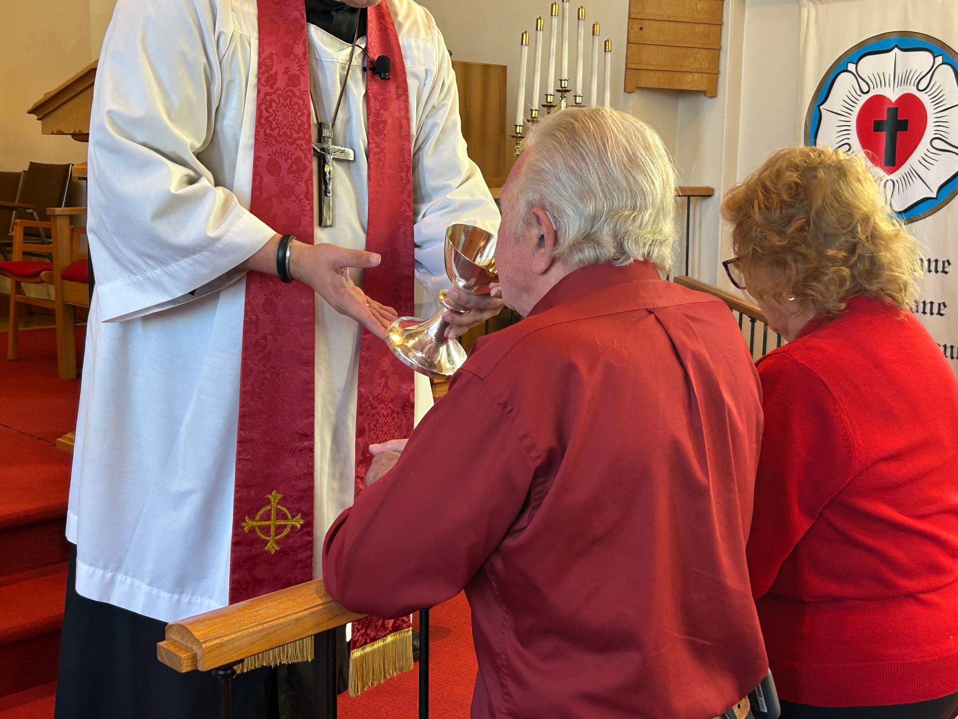A minister in white robes holds a silver chalice for a person kneeling to receive communion in a church.