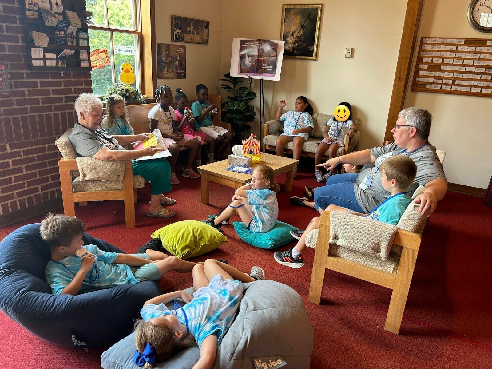 A group of children and adults sit in a cozy room with red carpeting, listening to a reading near an easel display.