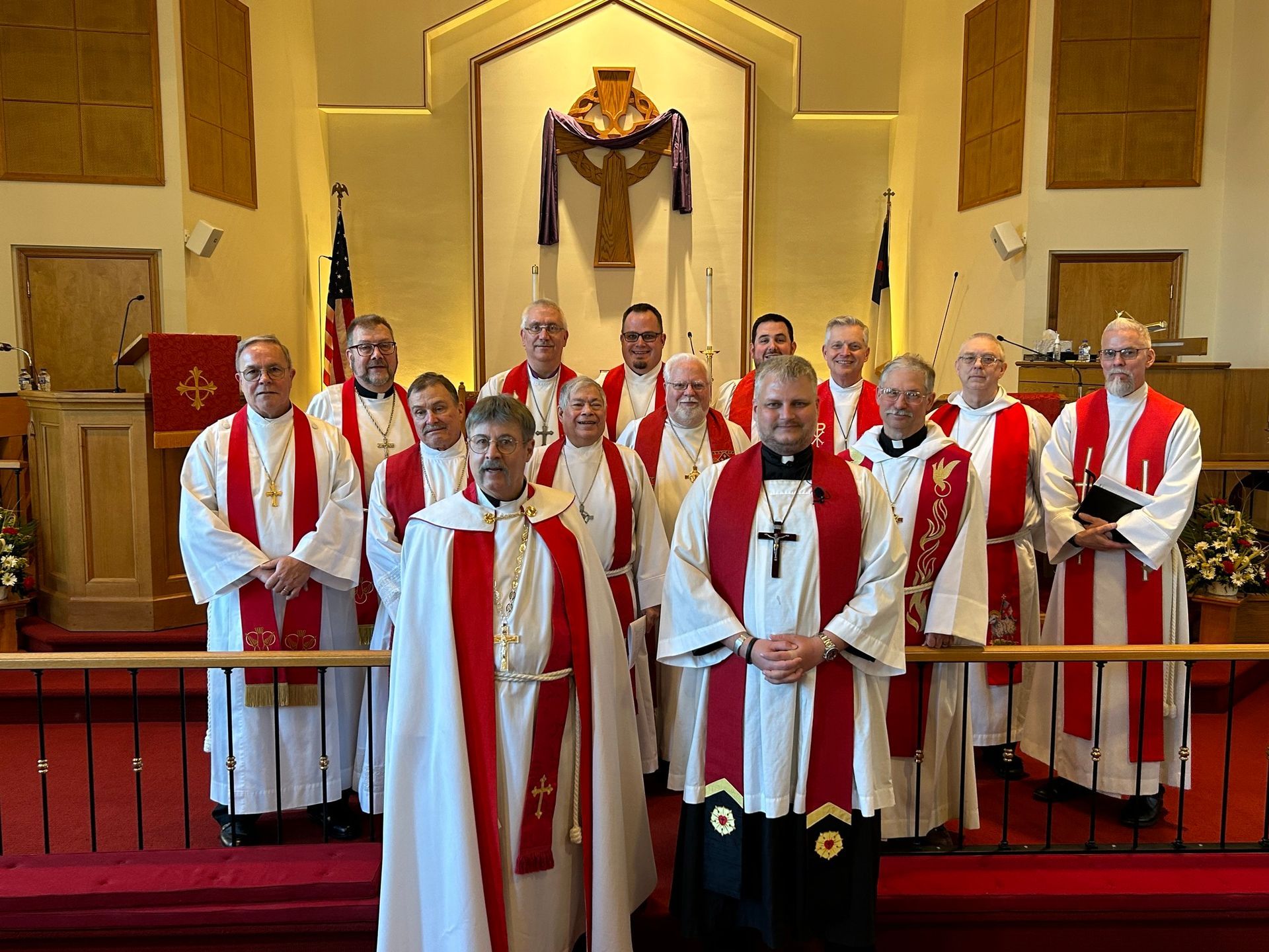 A group of clergy in white cassocks and red stoles pose for a photo in a church sanctuary.
