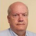 A bald man with a serious expression, wearing a light blue collared shirt against a plain, light-colored background.