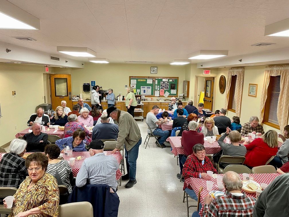 A crowded hall with people seated at tables with red checkered tablecloths, appearing to be at a community event.
