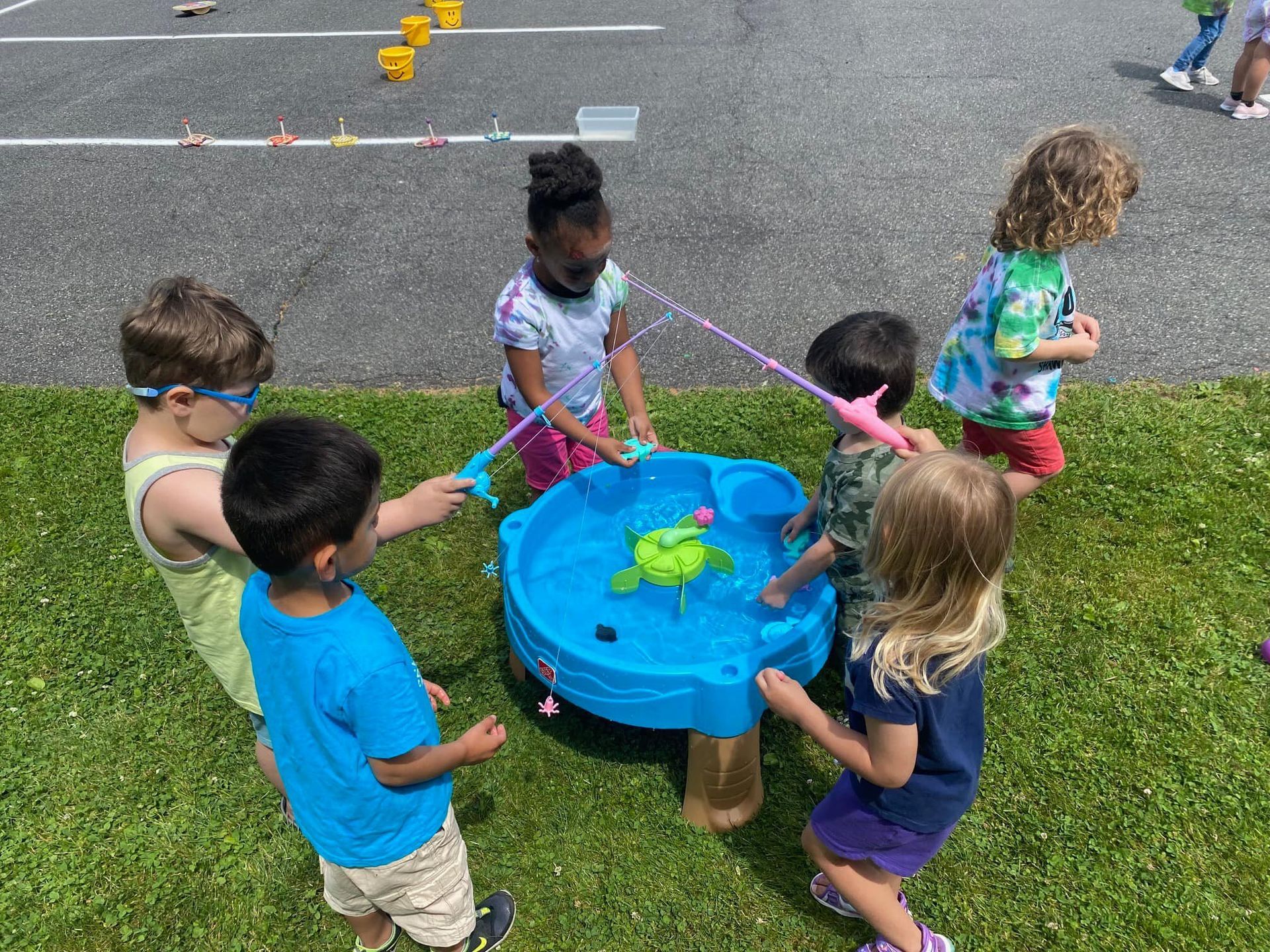 Five children stand around a blue circular fishing game toy on grass, using handheld fishing rods to catch items inside.