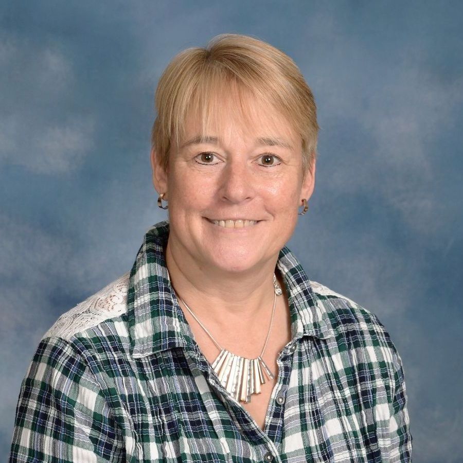A smiling person with short blonde hair wears a plaid shirt and a silver statement necklace against a blue background.