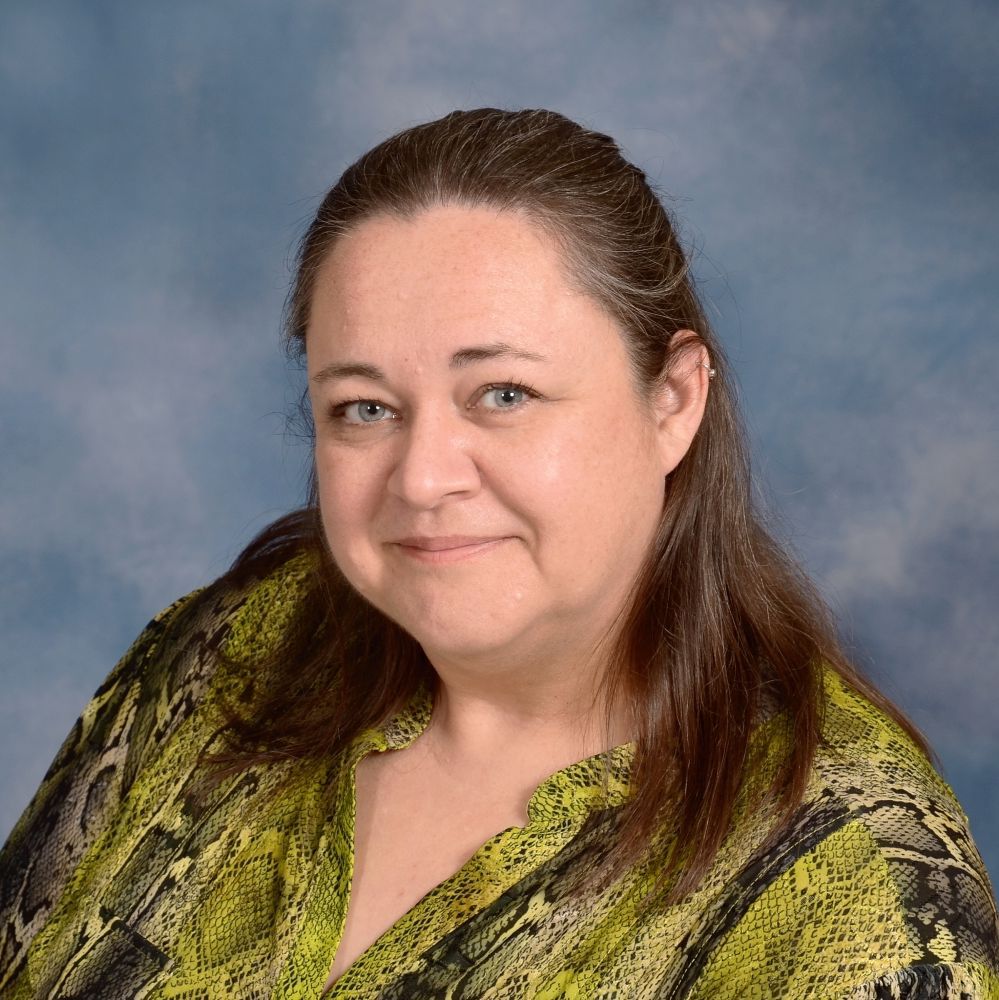 A person with shoulder-length brown hair, wearing a green snakeskin-patterned top, against a blue mottled background.