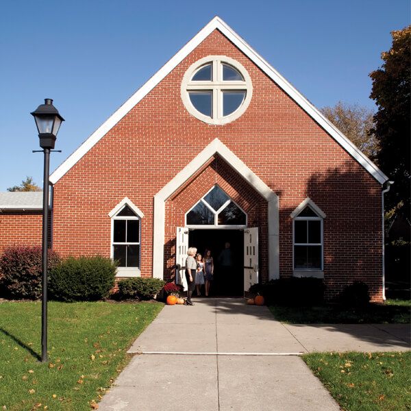 A brick church building with a cross-shaped window and a sidewalk leading to an open doorway with people inside.