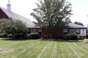 A red brick church building with a gray roof and a white steeple, surrounded by green trees and a mown lawn.