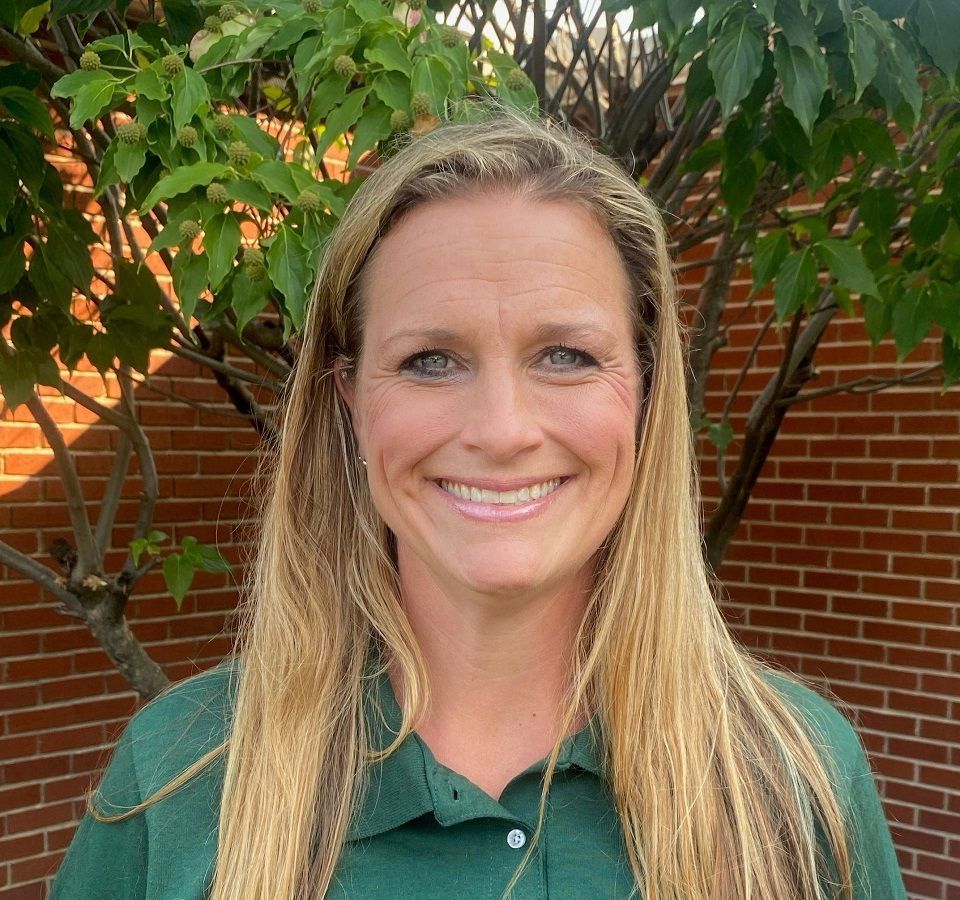A person with long blonde hair smiling, wearing a dark green polo shirt in front of a brick wall and leafy green tree.