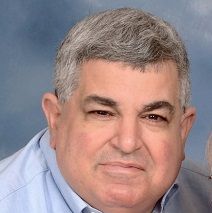 A person with short, graying hair wearing a blue button-down shirt against a soft blue background.