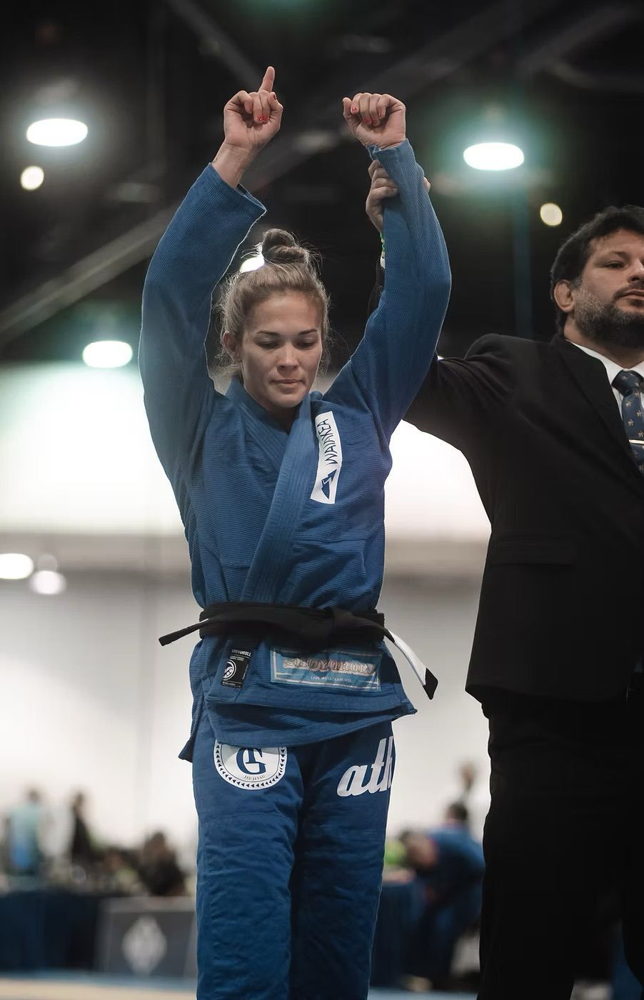 A competitor in a blue jiu-jitsu gi stands with arms raised in victory, held by a referee in a suit at a competition.
