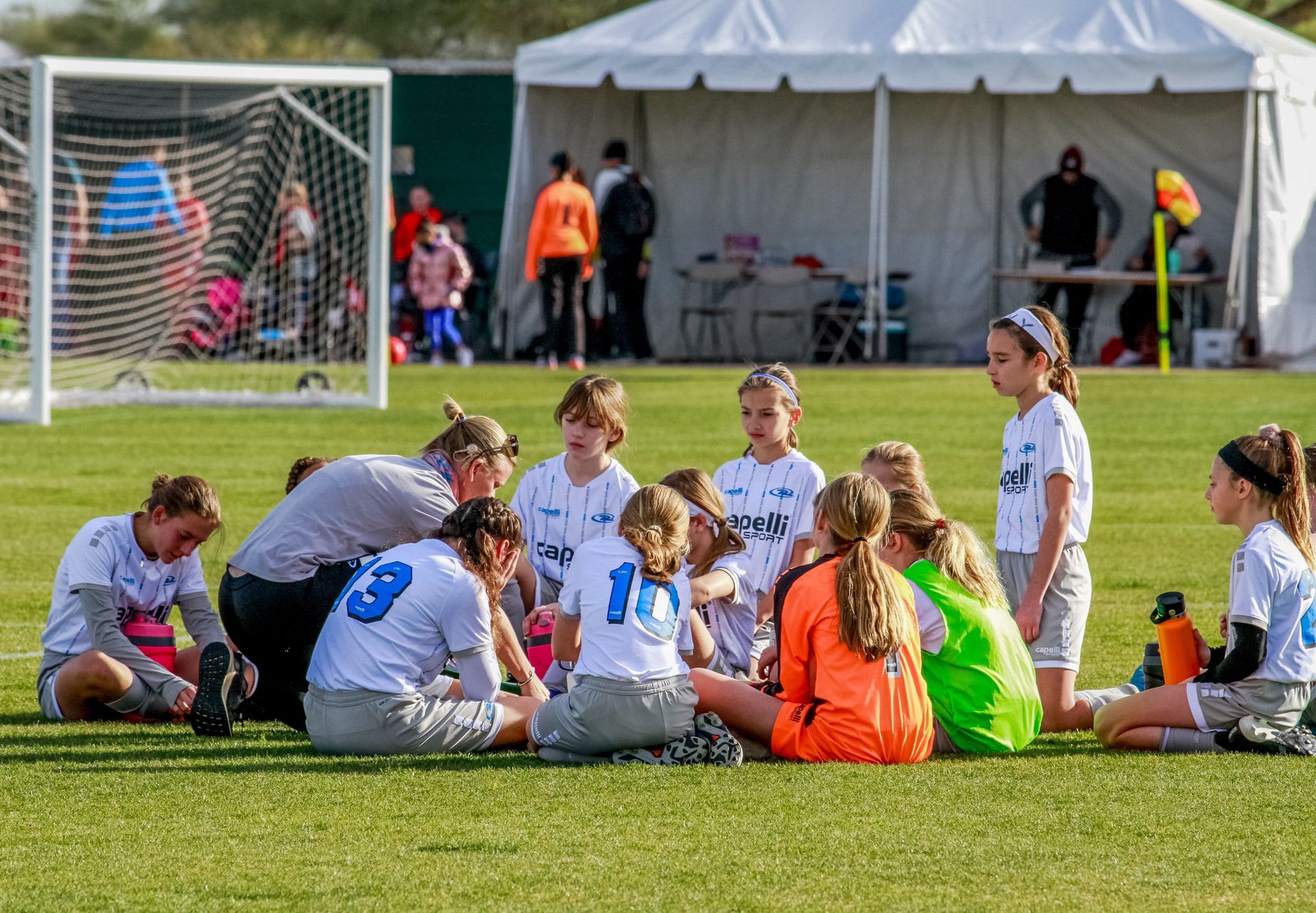 Youth soccer team sitting on grass near goal during a break, with coaches and a white tent in the background