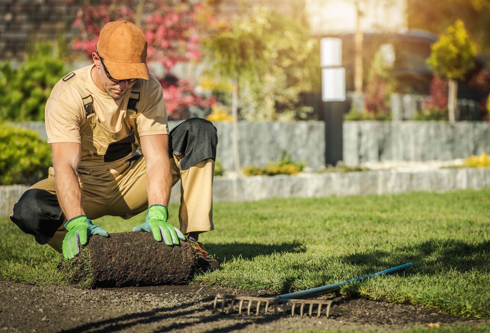 Gardener kneeling on grass, planting turf with green gloves and a rake nearby in a sunny yard