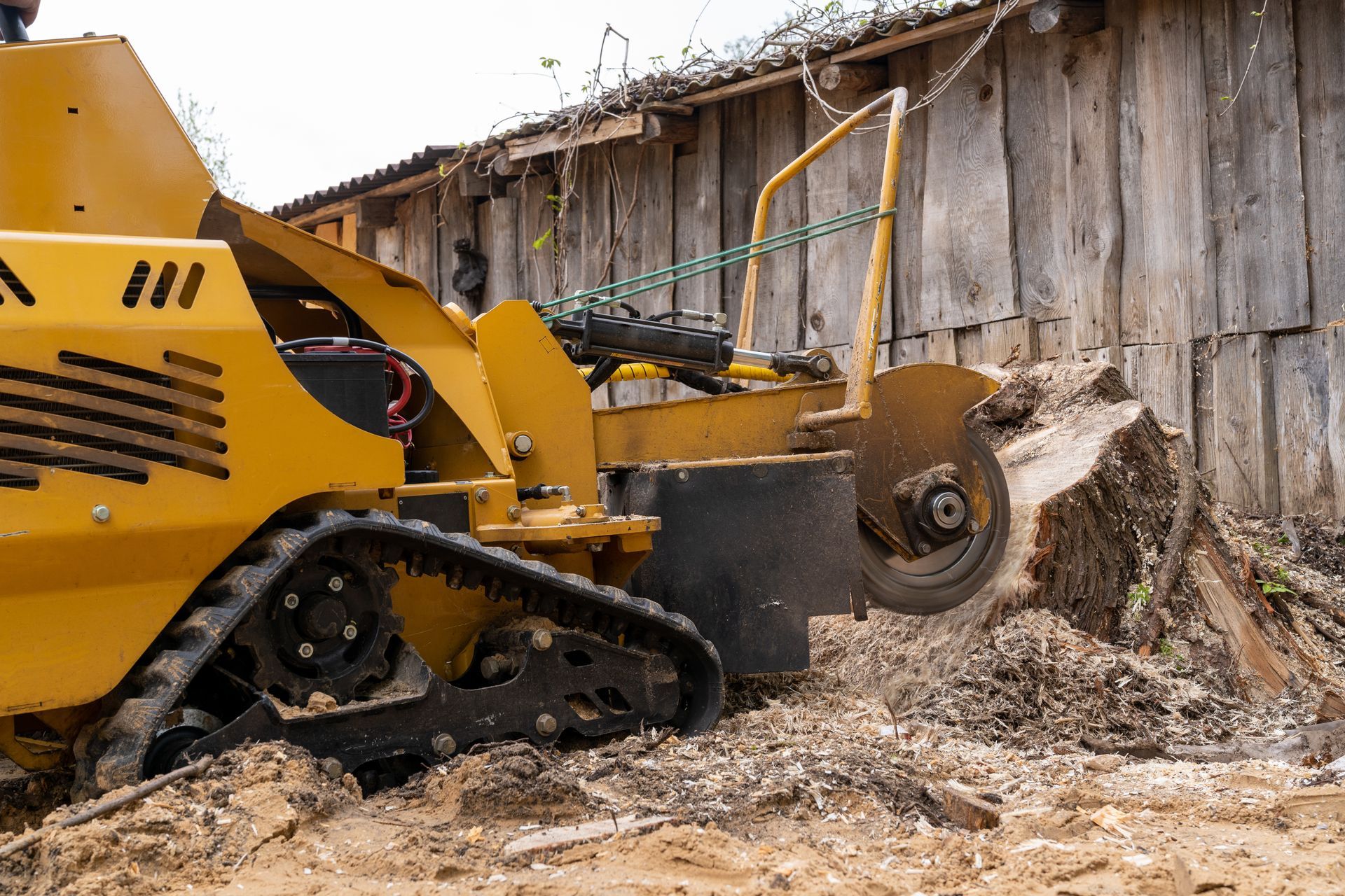 A yellow tractor is cutting a tree stump in front of a wooden building.