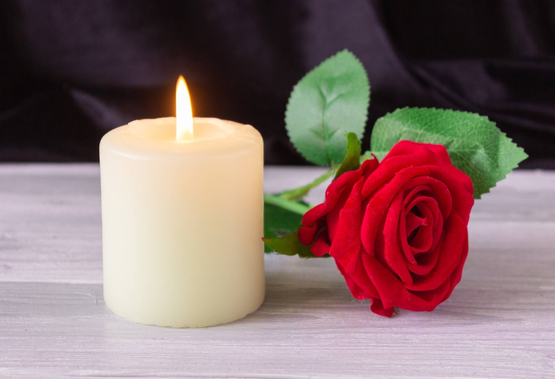 A lit white pillar candle sits next to a vibrant red rose on a light wooden surface against a dark background.