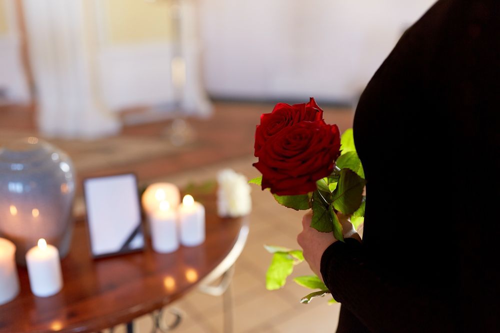 A person in black holds a red rose at a memorial service with a funeral urn, candles, and a framed photo on a table.