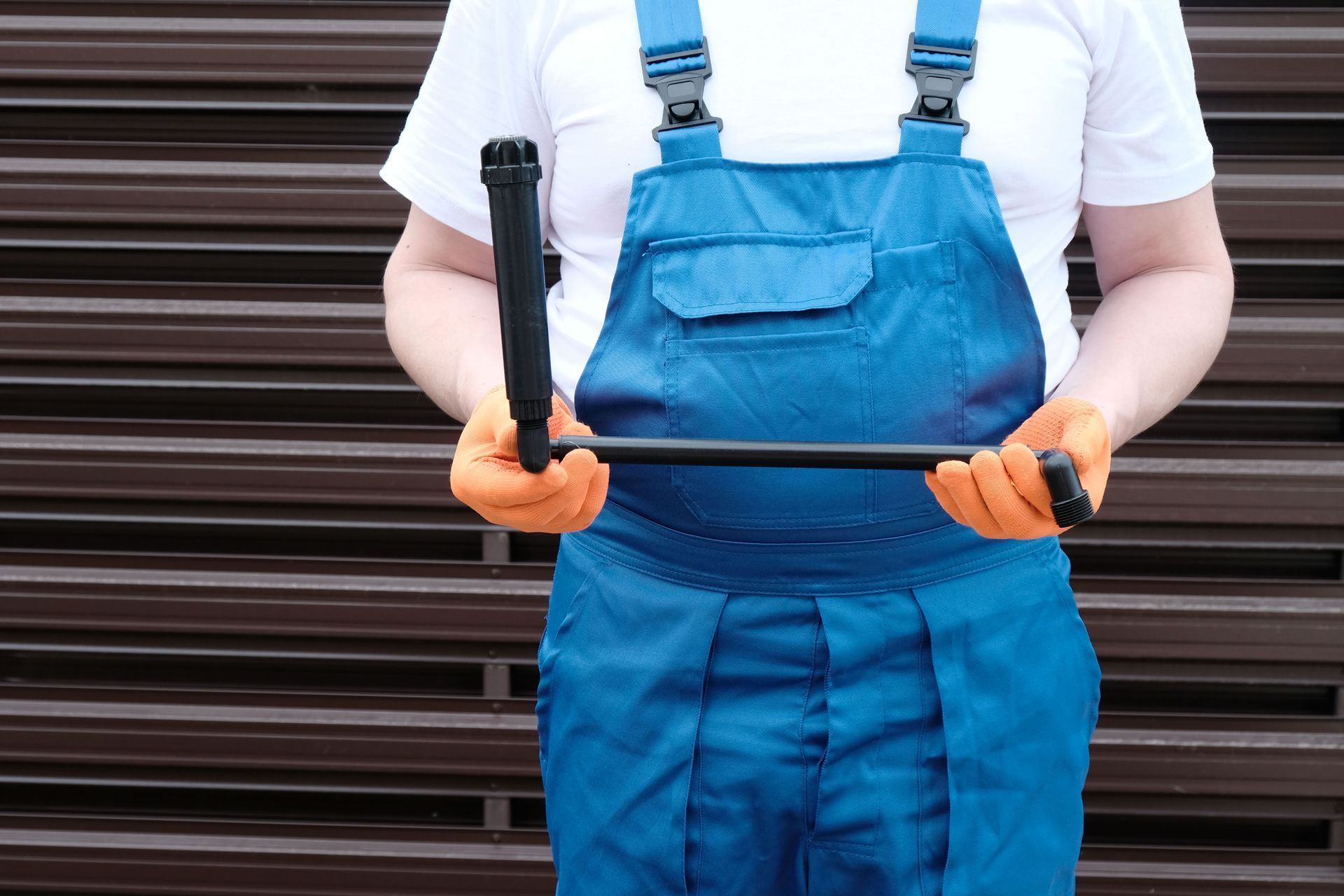 a person wearing blue overalls and orange gloves holding a black sprinkler horizontally.
