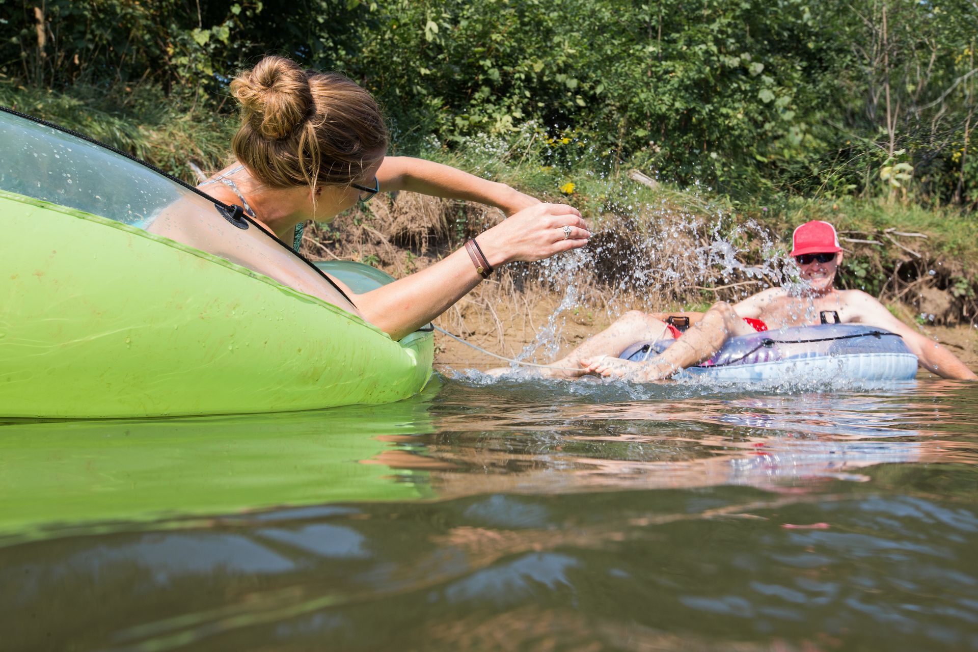 People splashing in a river from a green inflatable tube, with one wearing a red cap