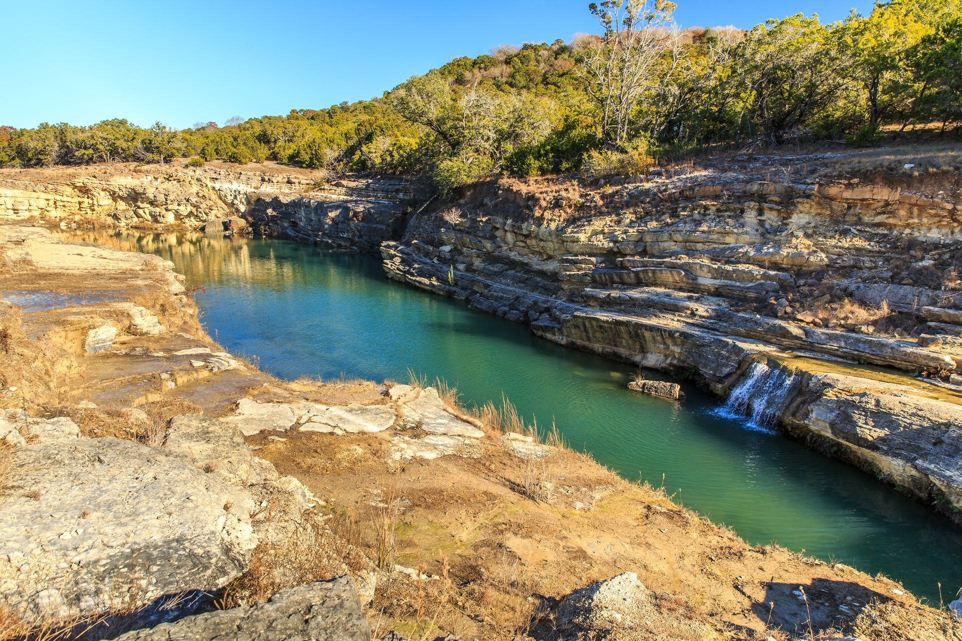 Rocky canyon with turquoise water and sunlit shrubs under a clear blue sky