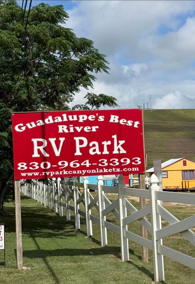 Red Guadalupe’s Best River RV Park sign beside white fence and green tree on a grassy hillside