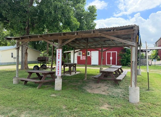 Outdoor picnic shelter with wooden tables, red building, and trees on a grassy lawn