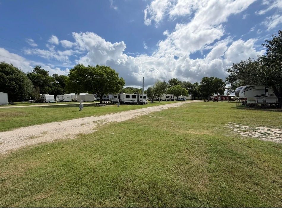 Sunny grassy field with a dirt path, parked RVs, and trees under a partly cloudy sky