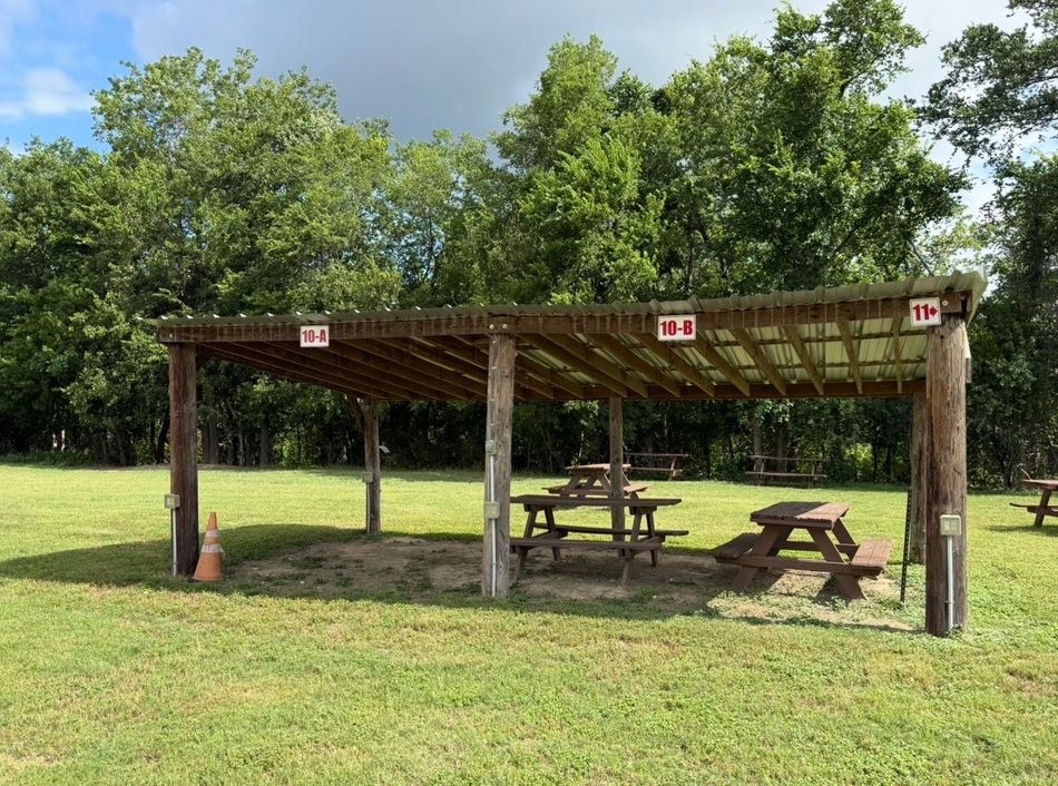 Wooden picnic shelter with tables in a grassy park under trees and a blue sky