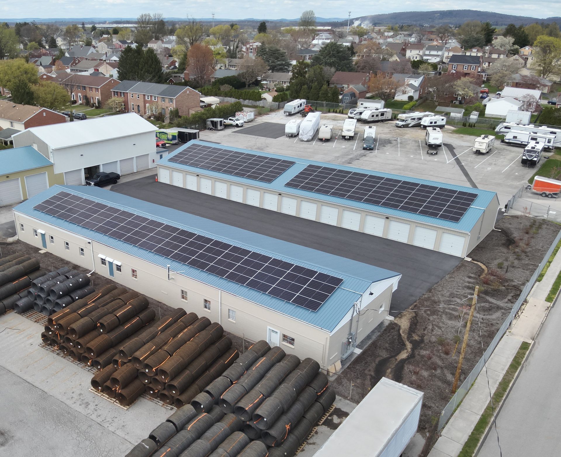 An aerial view of a self-storage facility with solar panels on the roofs, parked RVs, and stacks of pipes in the yard.