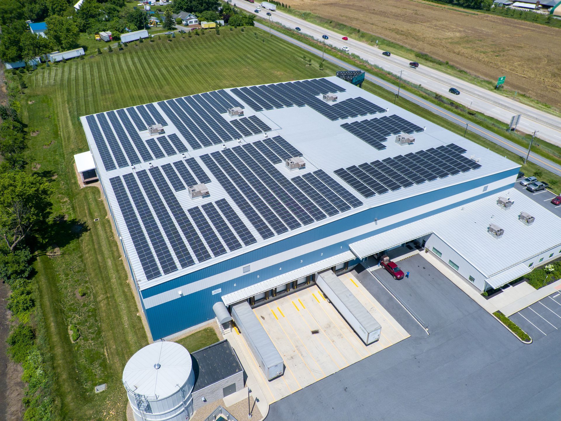 An aerial view of a warehouse with a large rooftop solar panel array, adjacent to a highway and a parking lot.