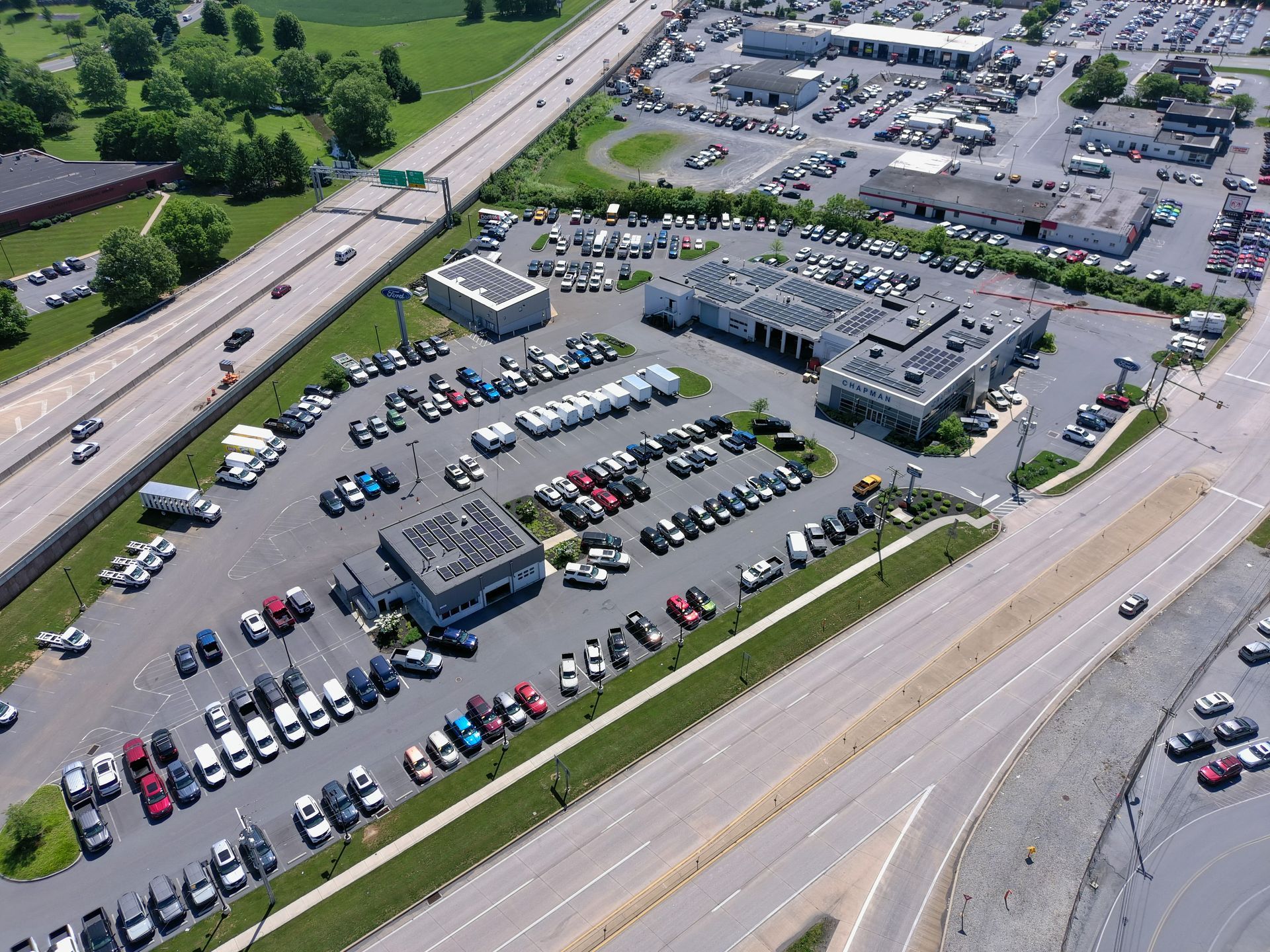An aerial view of a car dealership featuring a large parking lot full of vehicles, adjacent to a multi-lane highway.