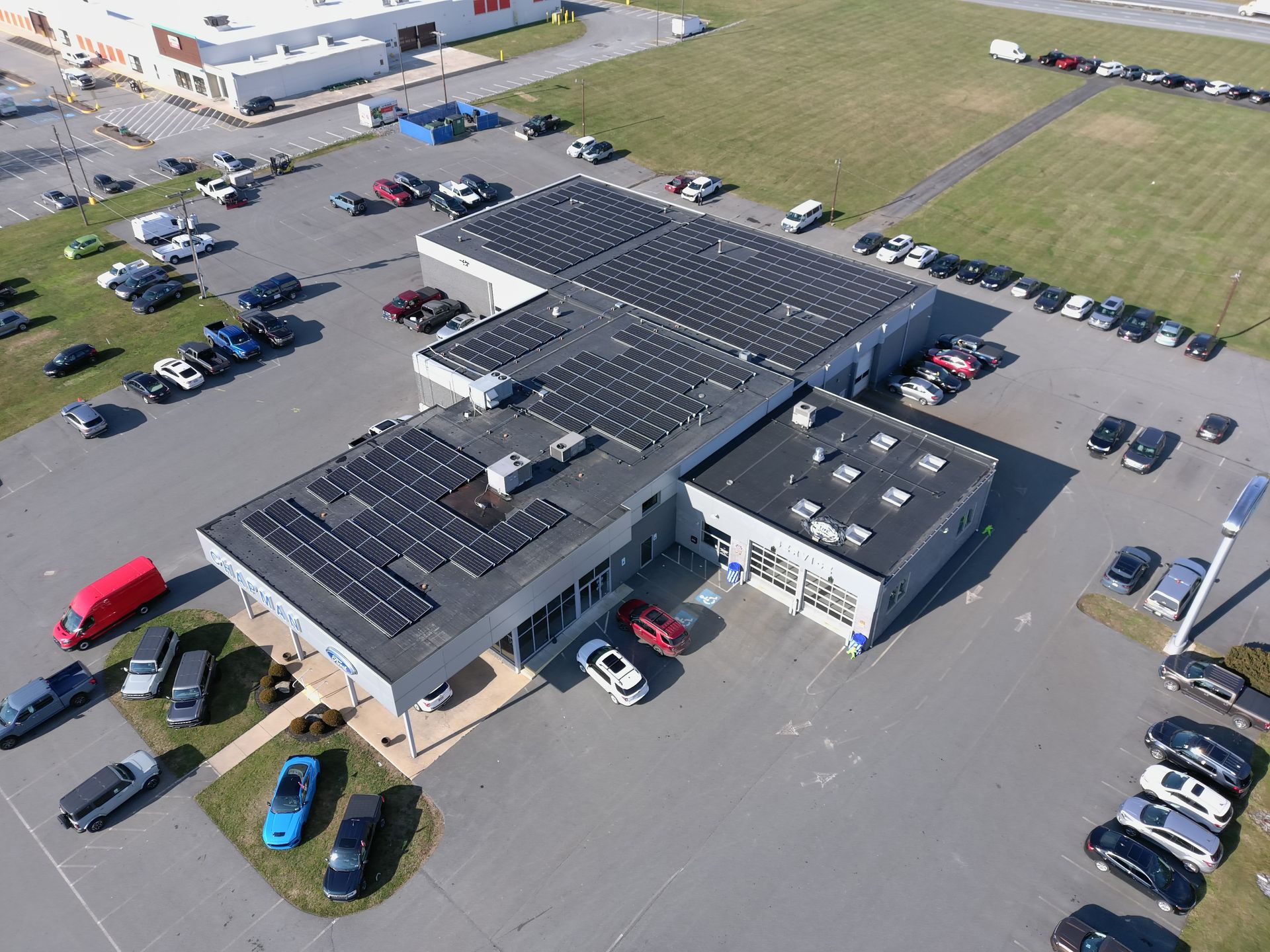 Aerial view of a commercial building with a large solar panel array on its roof, surrounded by a parking lot with cars.