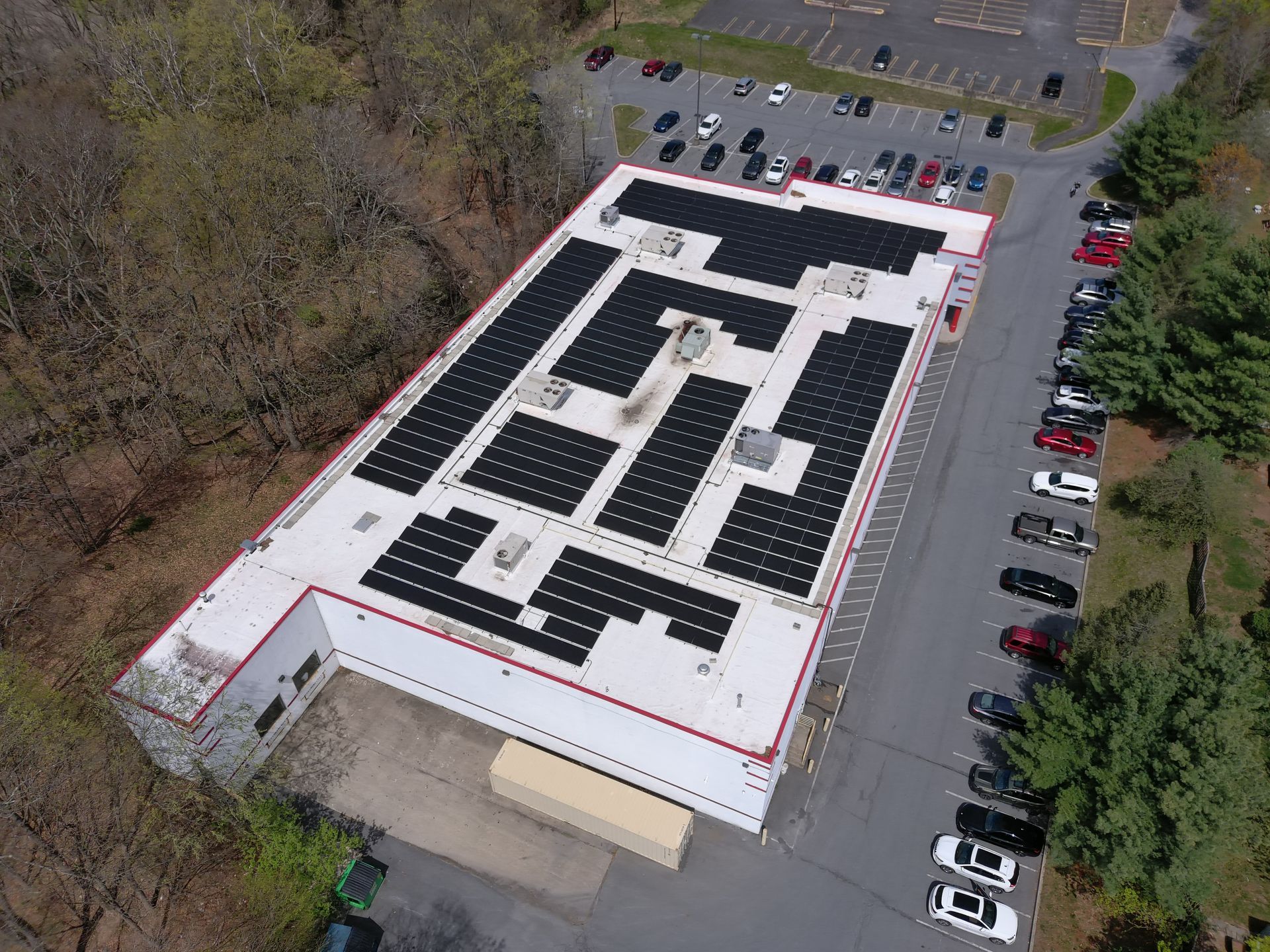 An aerial view of a flat-roofed commercial building with extensive solar panel arrays installed, surrounded by a parking lot.