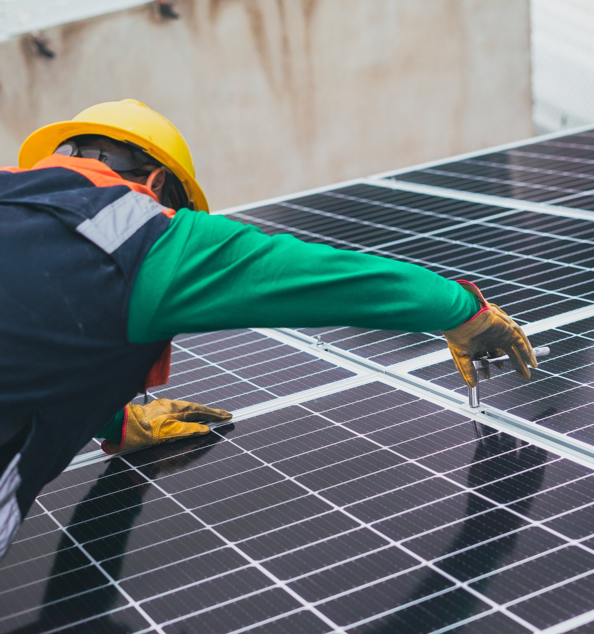 A worker in a yellow hard hat and gloves installs solar panels on a rooftop.