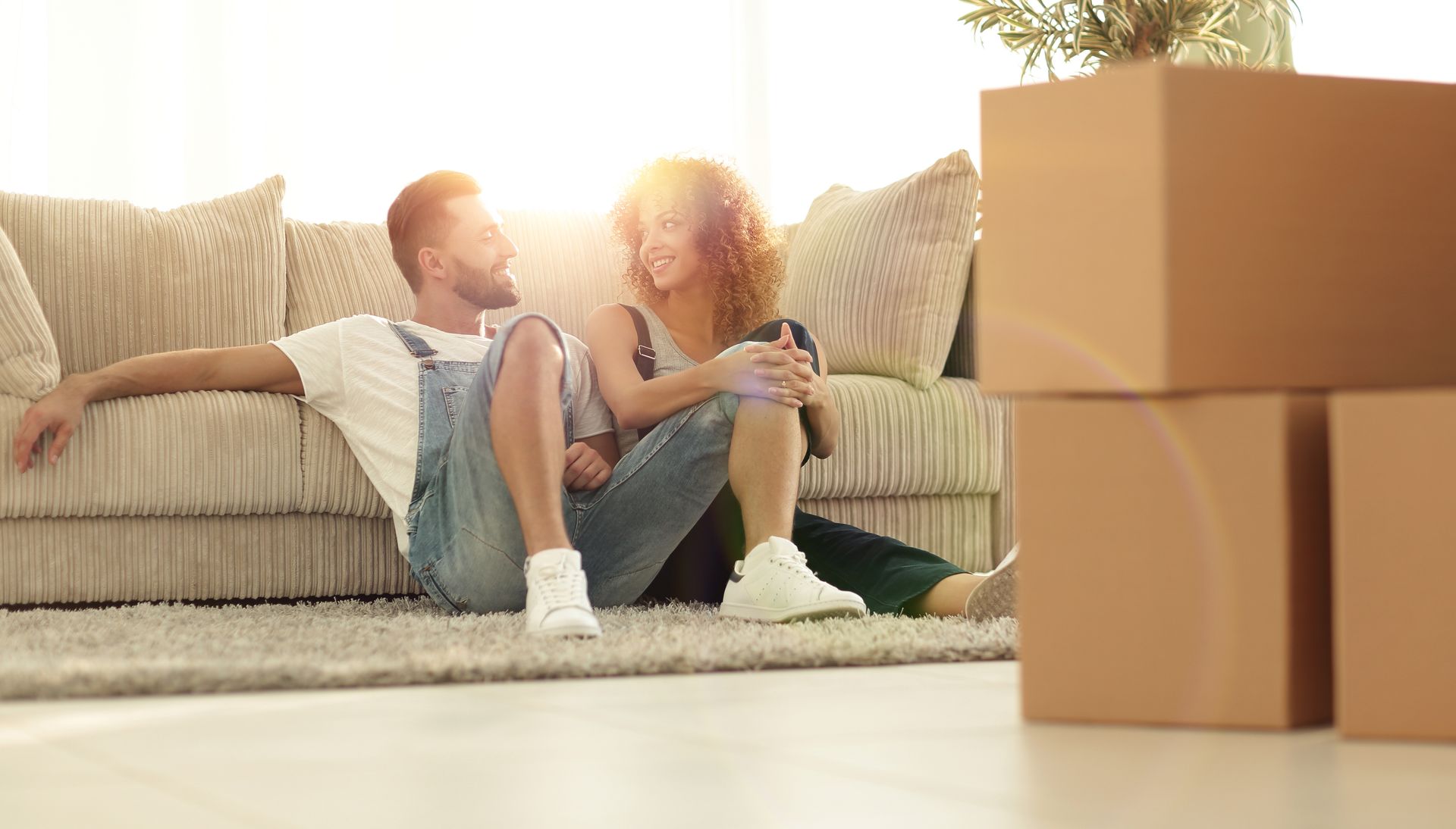A man and a woman are sitting on a couch in a living room surrounded by cardboard boxes.