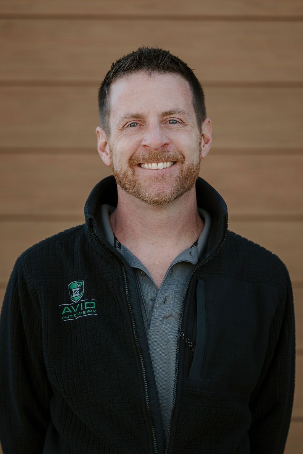 Smiling man in a black jacket standing against a wooden wall