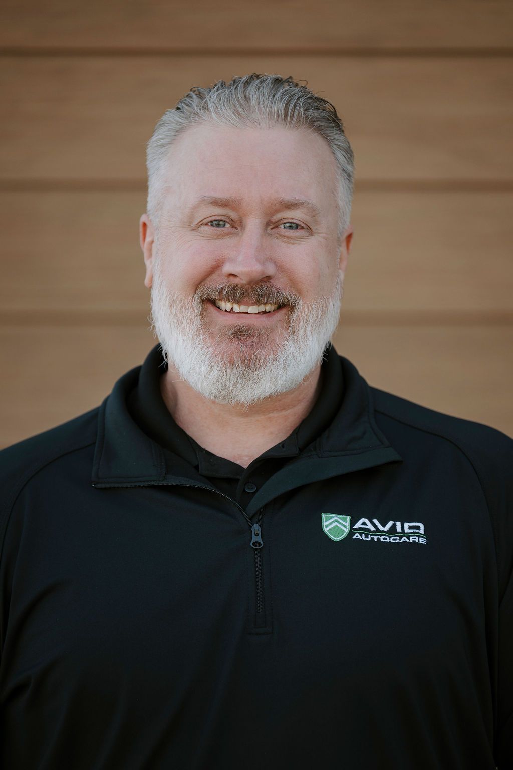 Portrait of a smiling man in a black AVID jacket against a wooden wall