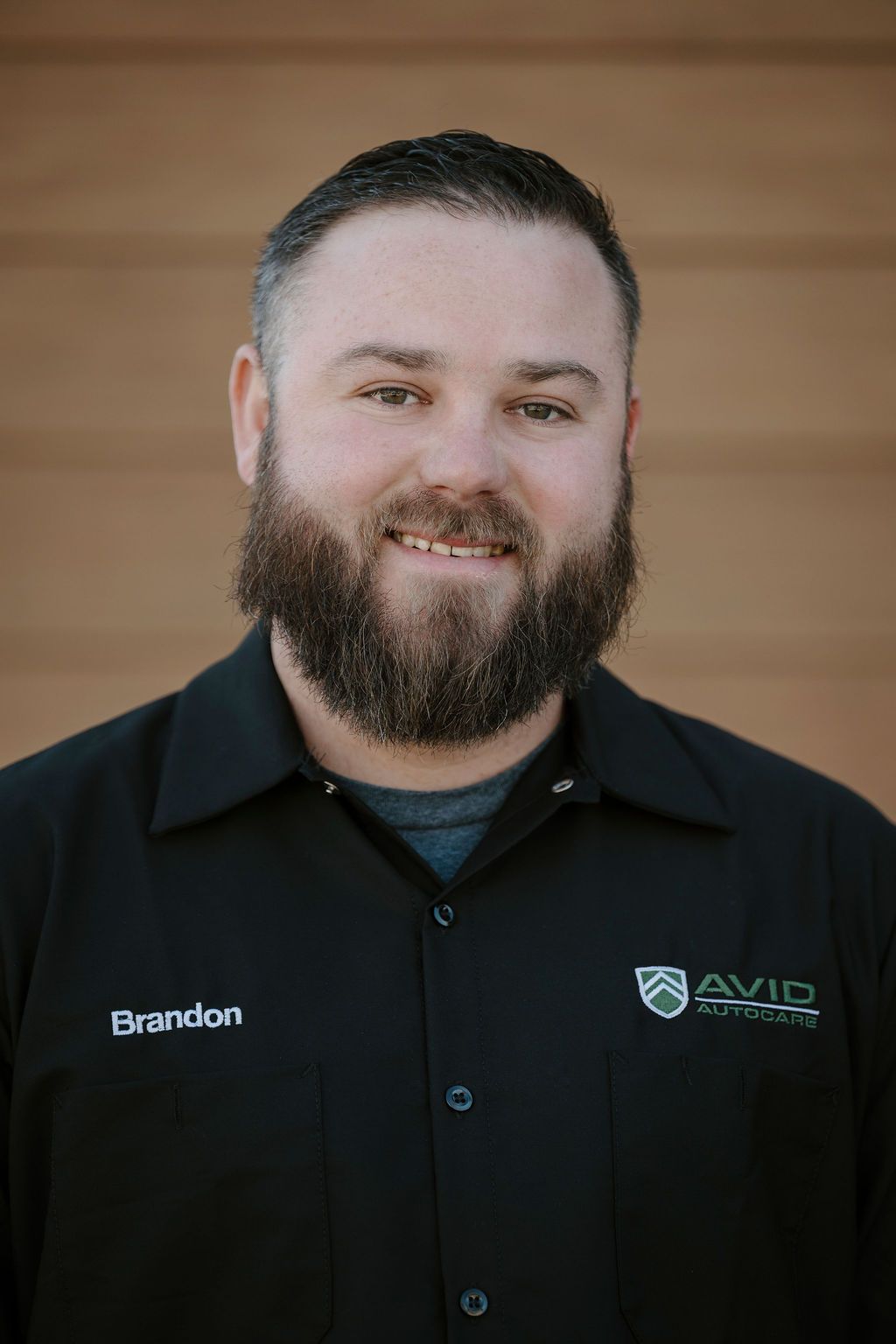 Portrait of a smiling man in a black collared shirt with logos against a wooden background
