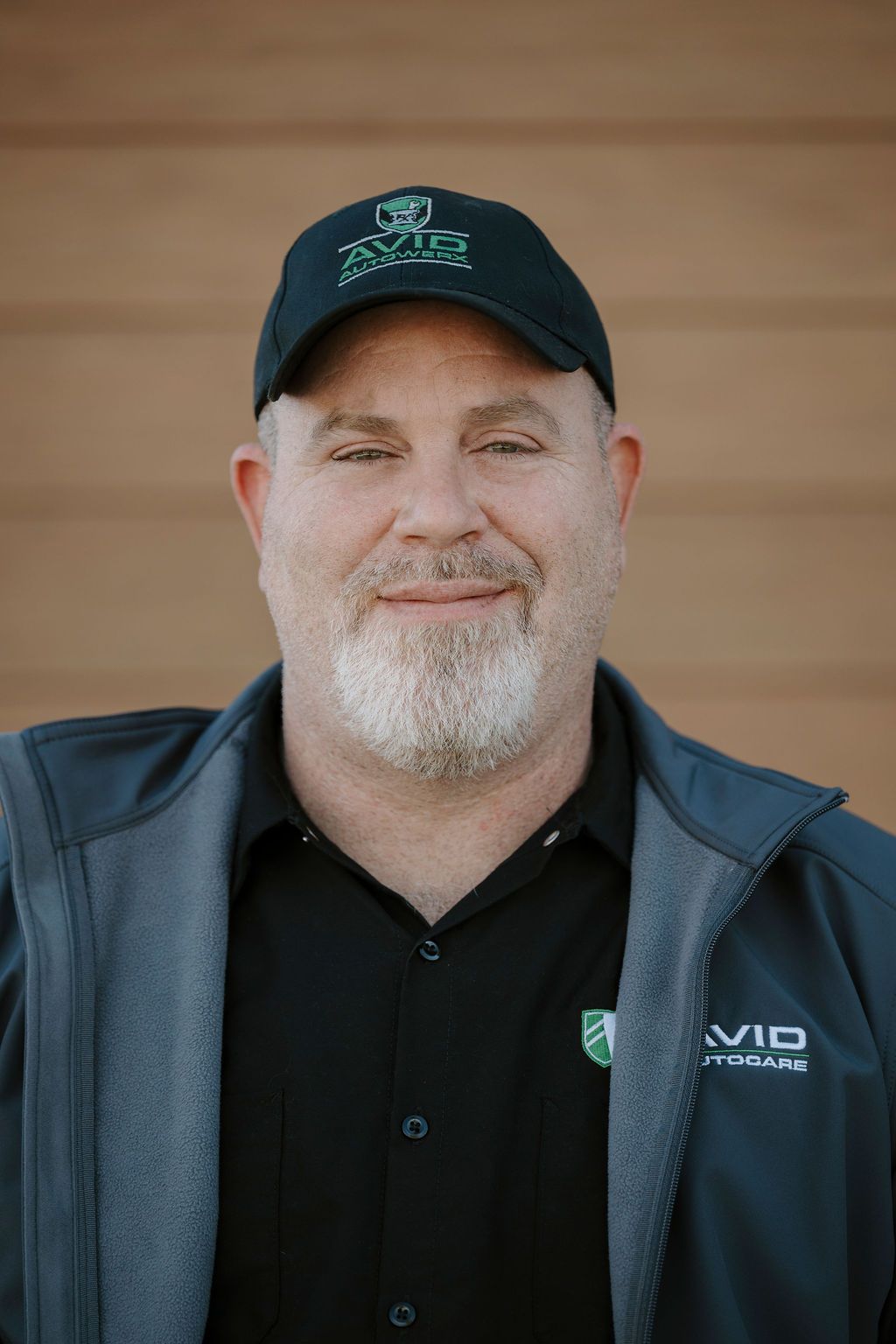 Smiling man in a black cap and jacket standing against a wooden wall