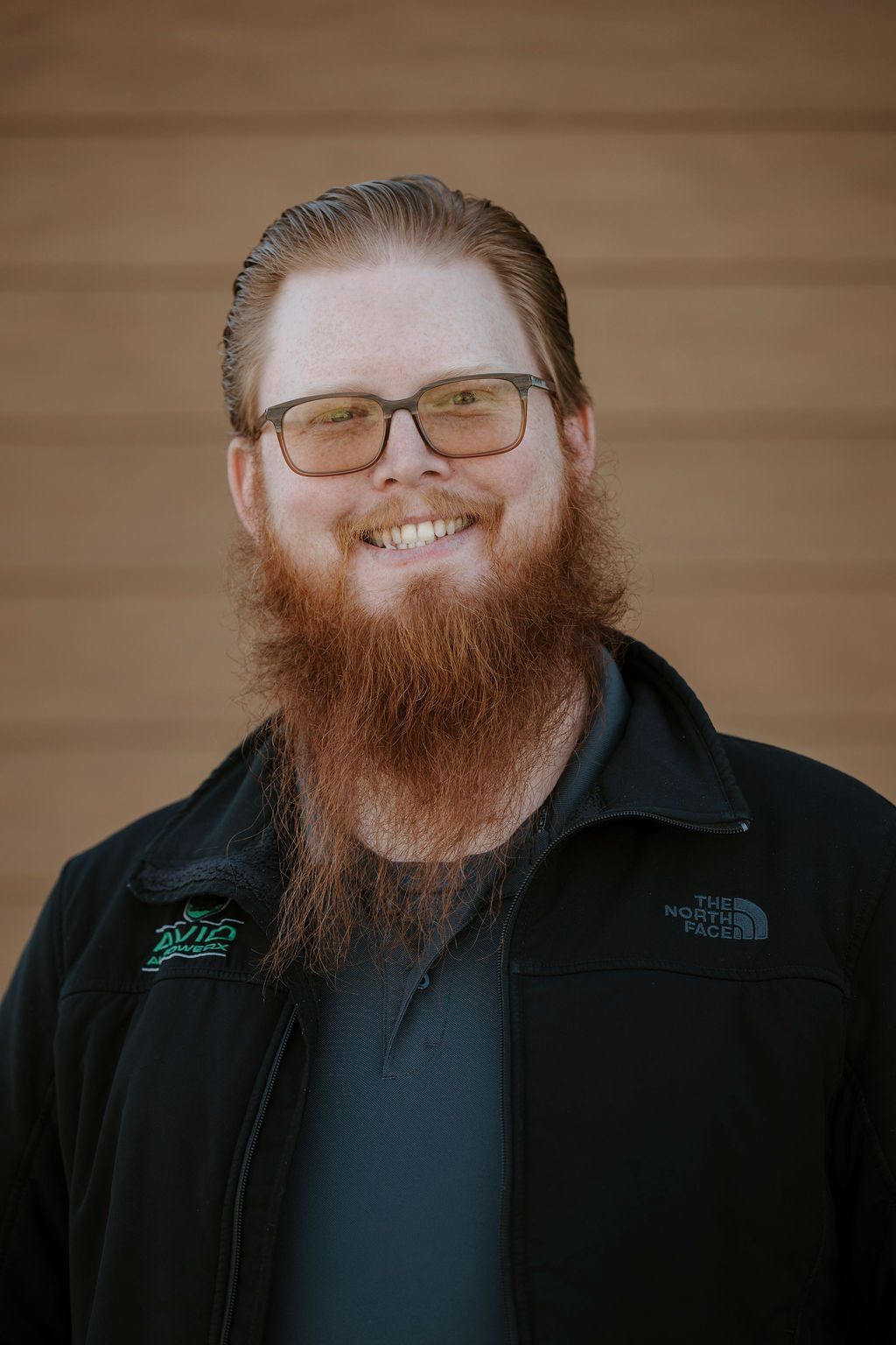 Smiling person with glasses and a long beard in a black jacket against a wooden wall