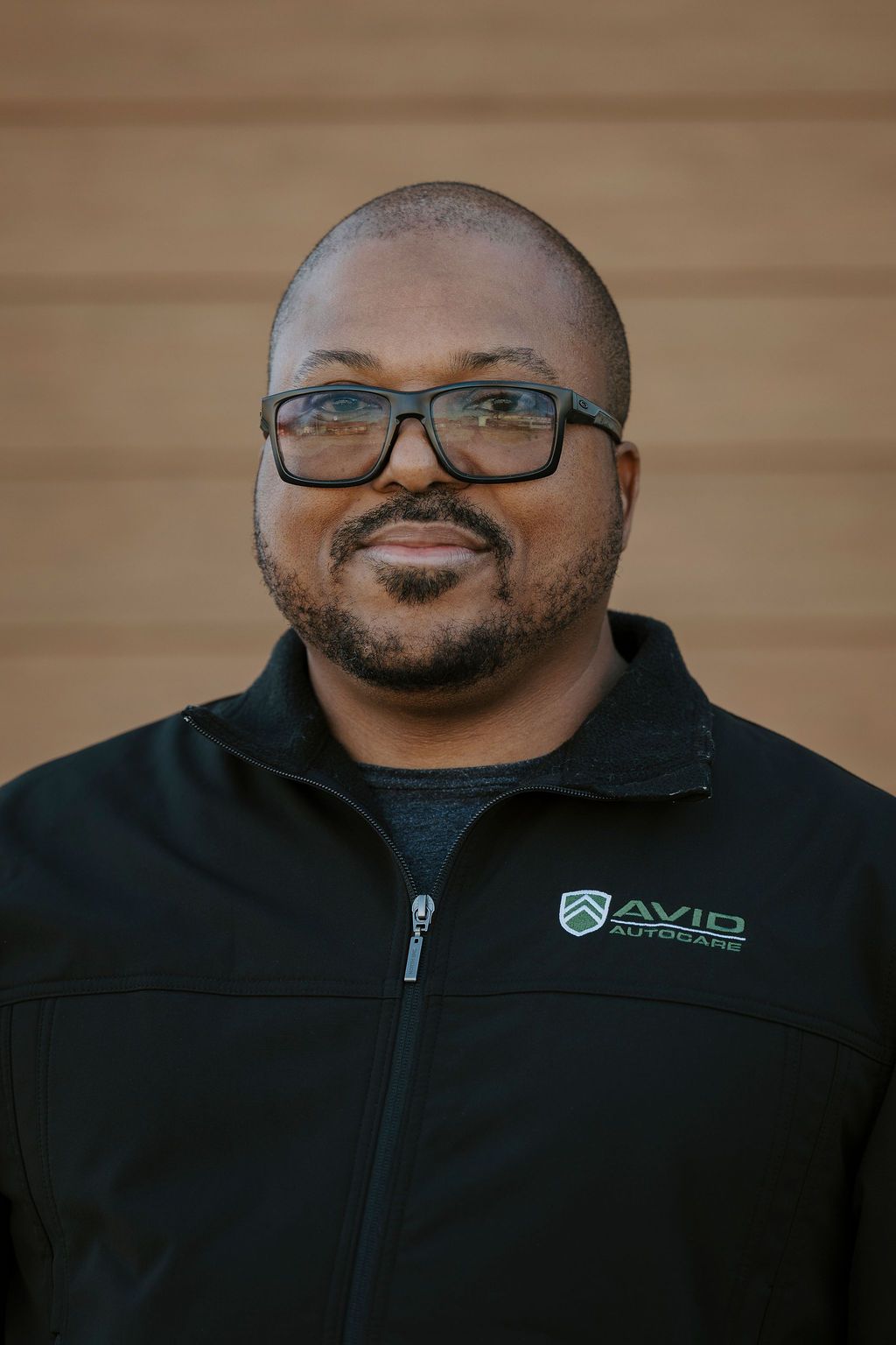 Smiling man in glasses wearing a black zip-up jacket with a logo, standing against a wood-paneled background