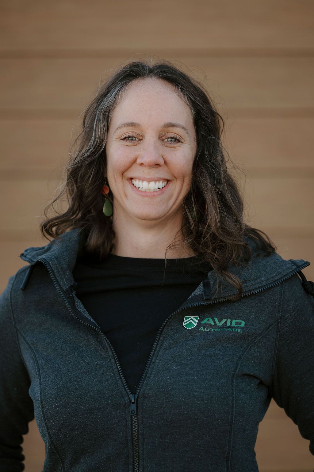 Smiling person in a dark jacket with a logo, standing against a wooden wall.