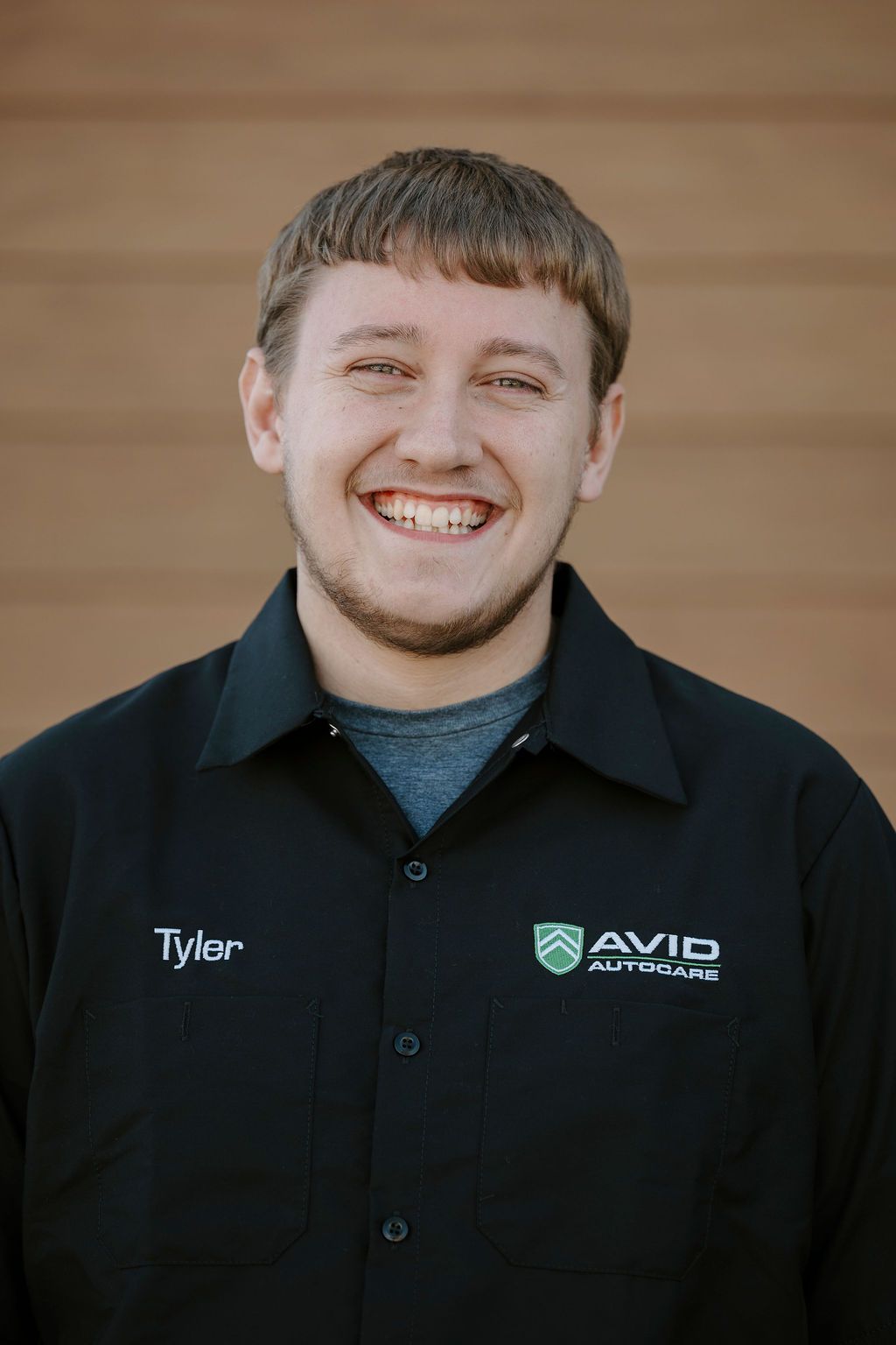 Smiling person in a black embroidered work shirt standing against a wooden wall