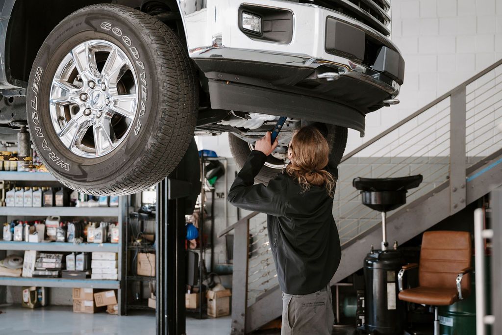 Mechanic working under a raised white truck in a garage with shelves of supplies nearby