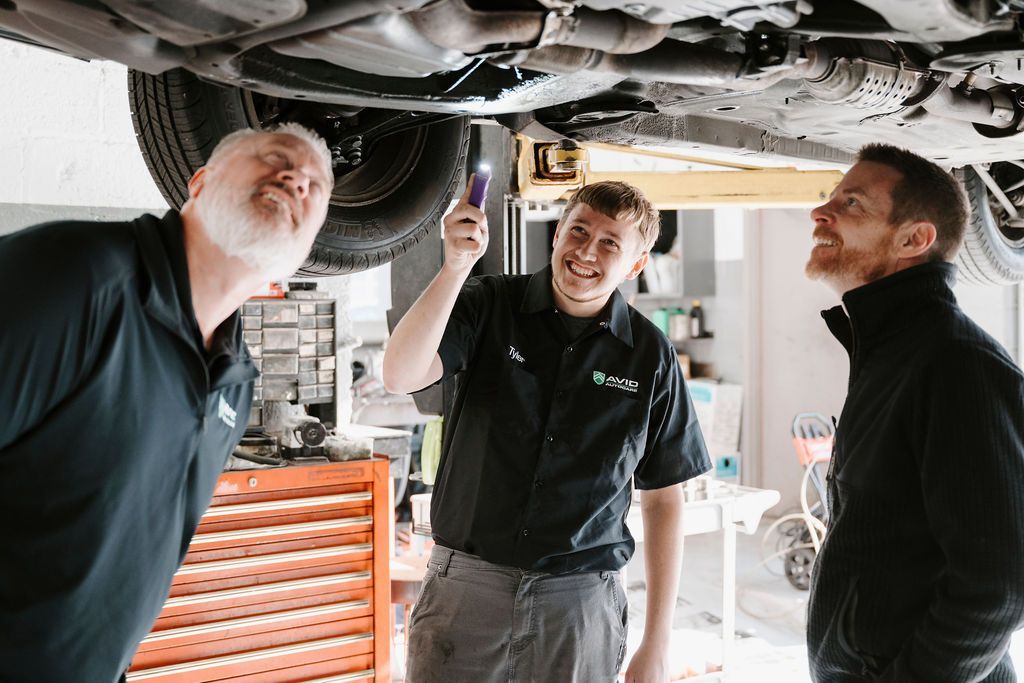 Three mechanics inspecting a car underside in a garage, one holding a tool and all smiling
