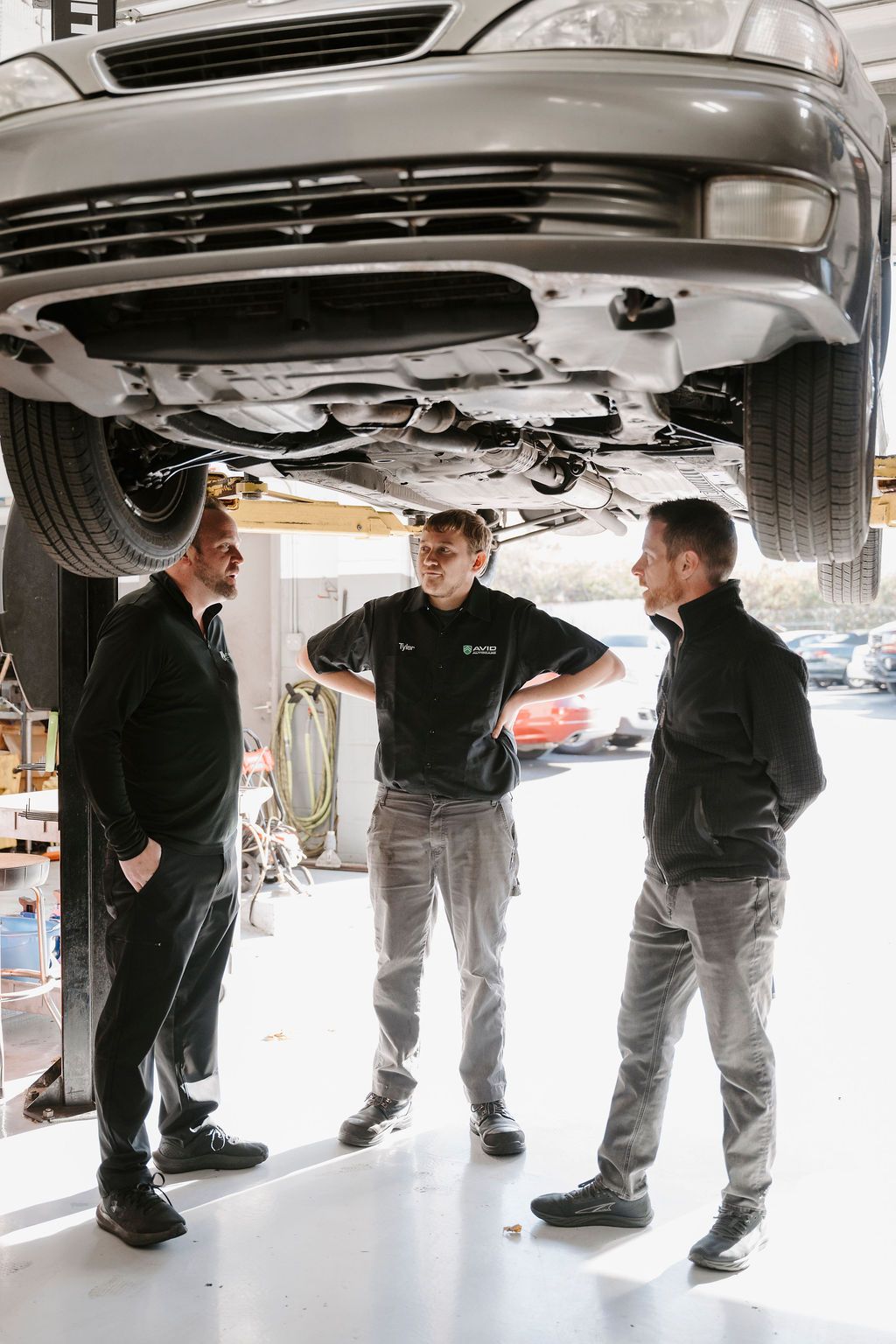Three people inspecting the underside of a car on a lift in an auto repair shop