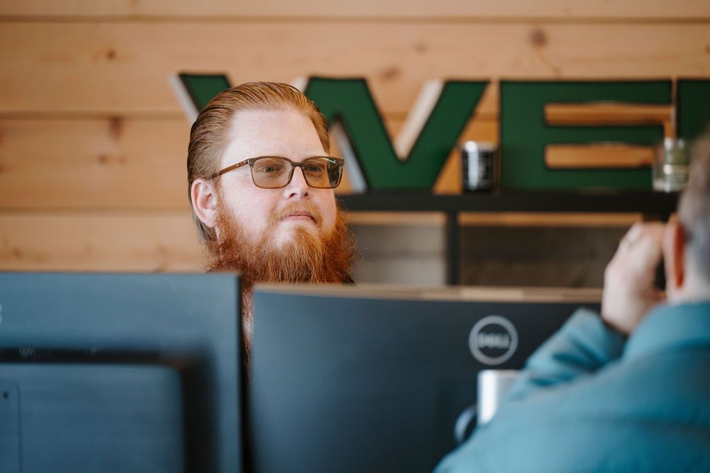 Bearded man in glasses behind office monitors, with a camera operator in the foreground.