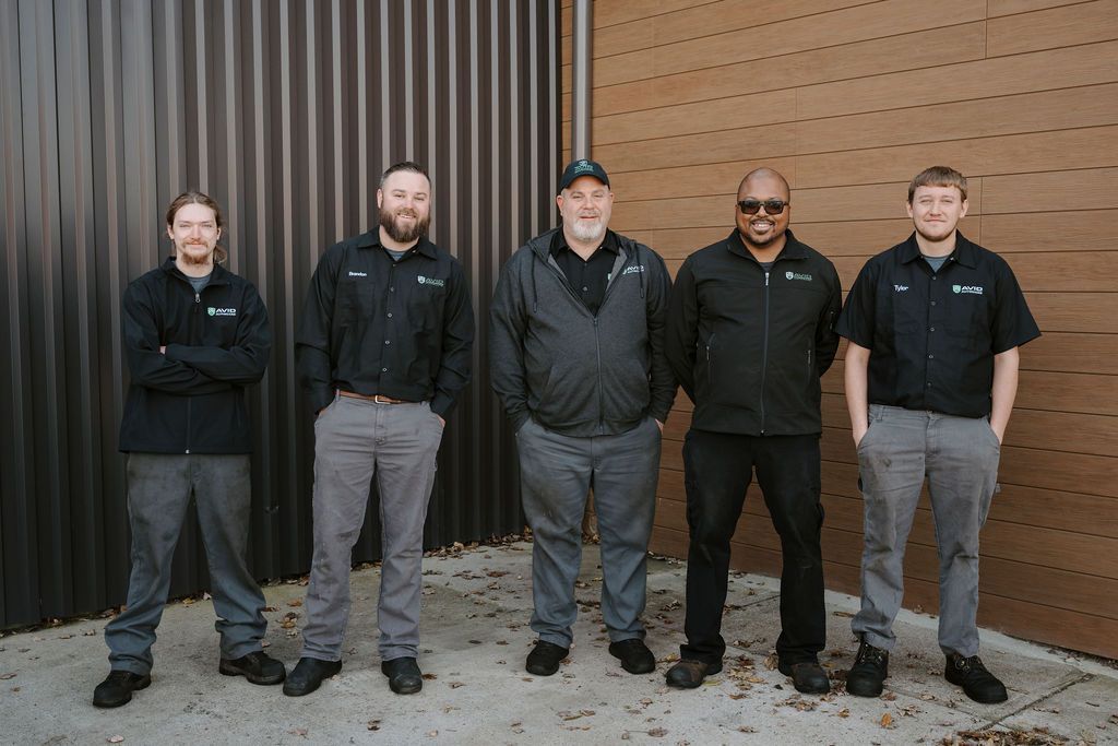 Five people standing outdoors against a corrugated metal wall, wearing dark jackets and jeans.