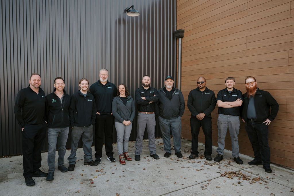 Group of nine people standing outdoors against a wooden and corrugated metal wall, posing for a photo.