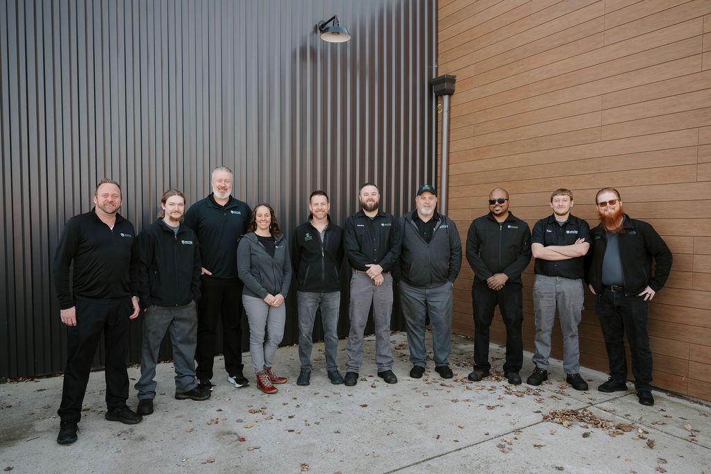 Nine people standing outdoors against a corrugated metal and wood wall, wearing dark jackets.
