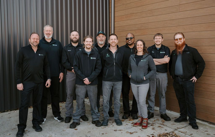 Nine people posing together outdoors in front of a brick wall, most wearing black jackets.